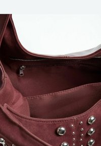 Unutrašnjost burgundske torbe s remenom sadrži odjeljak s patentnim zatvaračem, glatki platneni podstavljeni sloj, metalne zakovice na vanjskoj strani i mekanu teksturu.