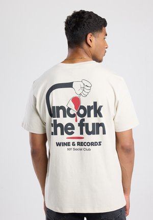 Crème katoenen t-shirt met een grafische print van een hand die wijn inschenkt, met vetgedrukte zwarte en rode tekst die "Uncork the Fun" leest.