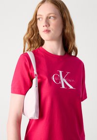 Camiseta de algodón fucsia con el logo blanco "CK", combinada con un pequeño bolso de hombro color crema que presenta una forma redondeada y un herraje metálico elegante.