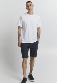T-shirt en coton blanc avec impression de logo, associé à un short cargo bleu marine avec de multiples poches. Baskets noires aux pieds nus, tatouages visibles.