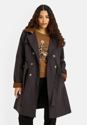 Mujer con cabello largo y rizado que lleva un abrigo marrón oscuro de doble botonadura sobre un suéter marrón con texto gráfico y pantalones negros.