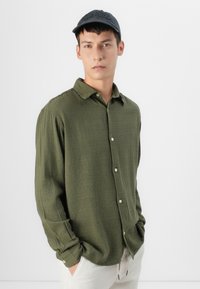 Green textured button-up shirt with a collar, featuring a relaxed fit. Paired with a dark cap and light-colored pants.