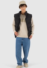 Zwarte puffer vest over een beige sweatshirt met de tekst "DARE", blauwe baggy jeans en beige sneakers; model draagt een lichtgekleurde bucket hat.