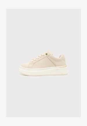 Beżowy niski sneaker z grubą białą podeszwą, sznurowaniem z przodu oraz wyściełanym kołnierzem z fakturą na obszarze pięty.