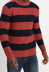 Knitted sweater with red and navy horizontal stripes, crew neck, ribbed texture, and cuffs. Worn over dark jeans.