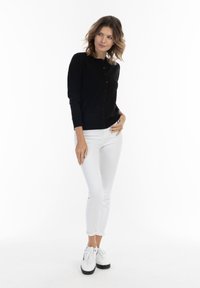 Cardigan negru cu închidere prin butoane, mâneci lungi și manșete cu dungi, asociat cu blugi albi și adidași cu talpă platformă. Fundal simplu.