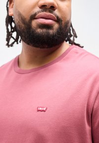 Άνδρας με γένια που φοράει ροζ πουκάμισο Levi's με εμφανές κόκκινο και λευκό λογότυπο στο στήθος, πάνω σε ουδέτερο φόντο.