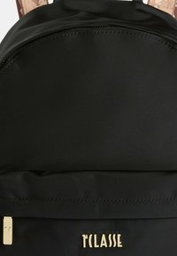 Czarny plecak z materiału o gładkiej teksturze, z zamkiem błyskawicznym na górze i złotym logo z przodu. Widoczne są brązowe paski w wzory.