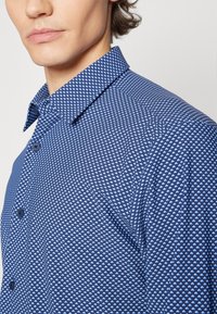 Jeune homme portant une chemise bleue à boutons avec un petit motif carré blanc, visible de l'épaule aux lèvres sur un fond uni.