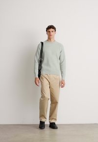 Jeune homme se tenant debout, portant un pull gris clair, un pantalon beige, des chaussures noires et un sac à bandoulière noir sur un fond blanc uni.