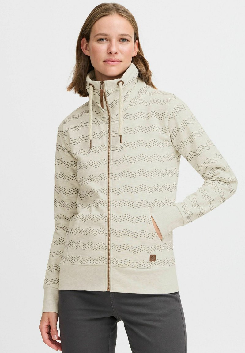 Veste zippée beige clair avec un col haut, présentant un motif en zigzag, des cordons de serrage et un petit logo marron sur la poche.