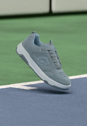 Grått atletisk joggesko med hvit såle som svever over blå og grønt tennisbanebane nær hvit grense linje.