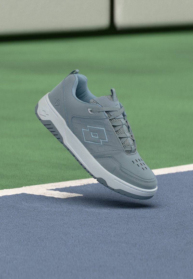 Γκρι αθλητικό παπούτσι με λευκή σόλα αιωρείται πάνω από την επιφάνεια γηπέδου τένις σε μπλε και πράσινο κοντά στην άσπρη γραμμή ορίου.