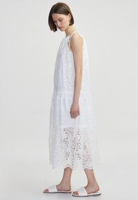 Vestido maxi blanco sin mangas con bordados florales, detalles de encaje, diseño con volantes y tirantes ajustables. Se lleva con sandalias blancas.