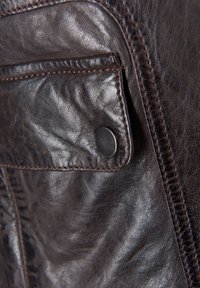 Kieszeń ciemno-brązowej skórzanej kurtki z klapką, zabezpieczoną okrągłym czarnym zatrzaskiem oraz widocznymi szwami na krawędziach.