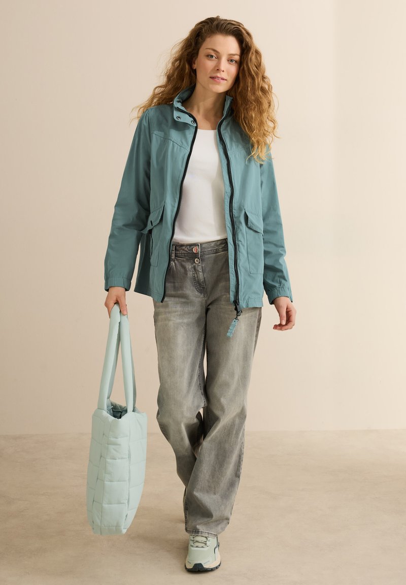 Veste légère bleu clair avec une fermeture éclair à l'avant et deux poches latérales, associée à un jean gris à jambes larges et un sac fourre-tout bleu clair assorti.