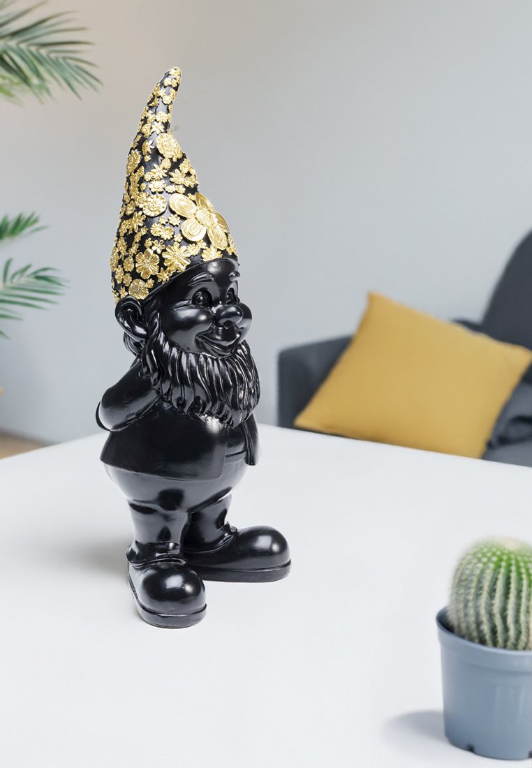 Figurine de gnome de jardin noire avec chapeau à motif floral doré sur table blanche, coussin jaune sur canapé gris et petit cactus en pot à proximité.