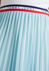 Lichtblauwe geplooide rok met een textured afwerking, met een witte tailleband met rode en marineblauwe accenten en de tekst "TOMMY JEANS".