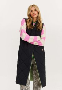 Schwarwest gesteppter langer Weste mit grünem Innenfutter, kombiniert mit einem rosa-weißen gestreiften Pullover und gemusterten hellen Hosen.