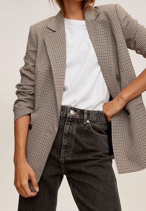 Kvinde iført en ternet blazer over en hvid t-shirt og sorte, højtaljede jeans, hånd i lommen, afslappet pose mod ensfarvet baggrund.