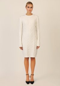 Weißes Strickkleid mit Rautenmuster, rundem Halsausschnitt, langen Ärmeln und geripptem Saum, kombiniert mit schwarzen High Heels.