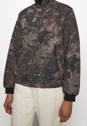 Personne portant une veste zippée à motif camouflage avec ourlet et poignets élastiques sur un pantalon clair, mains détendues sur les côtés.