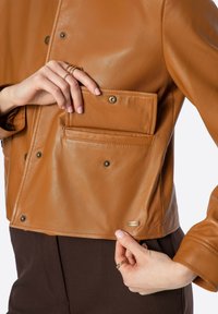 Giacca in pelle marrone con finitura liscia, dotata di una tasca frontale con chiusura a pressione e un marchio metallico vicino all'orlo.