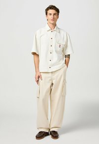 Crème shirt met korte mouwen, knopen en een zak, gecombineerd met beige cargobroek met brede pijpen, zijzakken en een relaxte pasvorm. Brown schoenen.