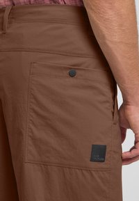 Bruine cargoshorts met een grote zijzak, een gladde textuur en een zwart logo-opdruk. Voorzien van een drukknoopsluiting op de zak.