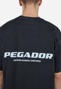 Svart bomullst-skjorte med avslappet passform, med stor hvit tekst "PEGADOR" og "CERTIFIED AUTHENTIC STREETWEAR" på baksiden.