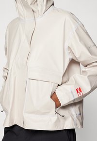 Veste imperméable beige avec capuche, dotée de deux poches avant, d'éléments réfléchissants et de cordons ajustables à l'ourlet et aux poignets.