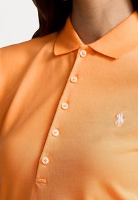 Orange polotröja i texturerad tyg med klassisk krage, sex knappar och en vit broderad logotyp på vänster bröst.