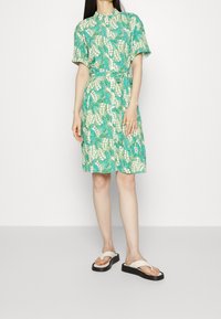 Kurzärmliges Kleid in Grün und Creme mit botanischem Muster, tailliert mit Gürtel und einem Kragen; kombiniert mit flachen Sandalen.