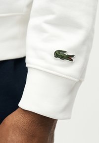Manchette blanche à manches longues avec un petit logo crocodile vert brodé près du poignet sur une personne à la peau foncée.