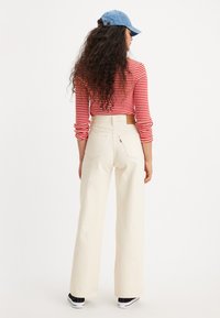 Pantalons larges crème avec taille haute et poches arrière, associés à un haut à manches longues rayé rouge et blanc et une casquette de baseball bleue.
