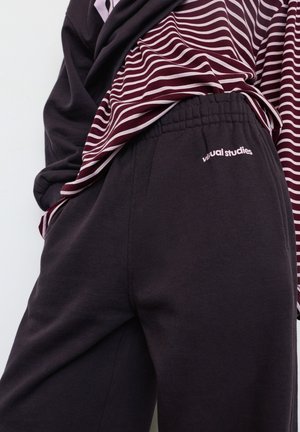 Zwarte joggingbroek met een elastische tailleband, voorzien van een klein logo met de tekst "visual studies". Gedragen met een bordeauxrode en wit gestreepte top.