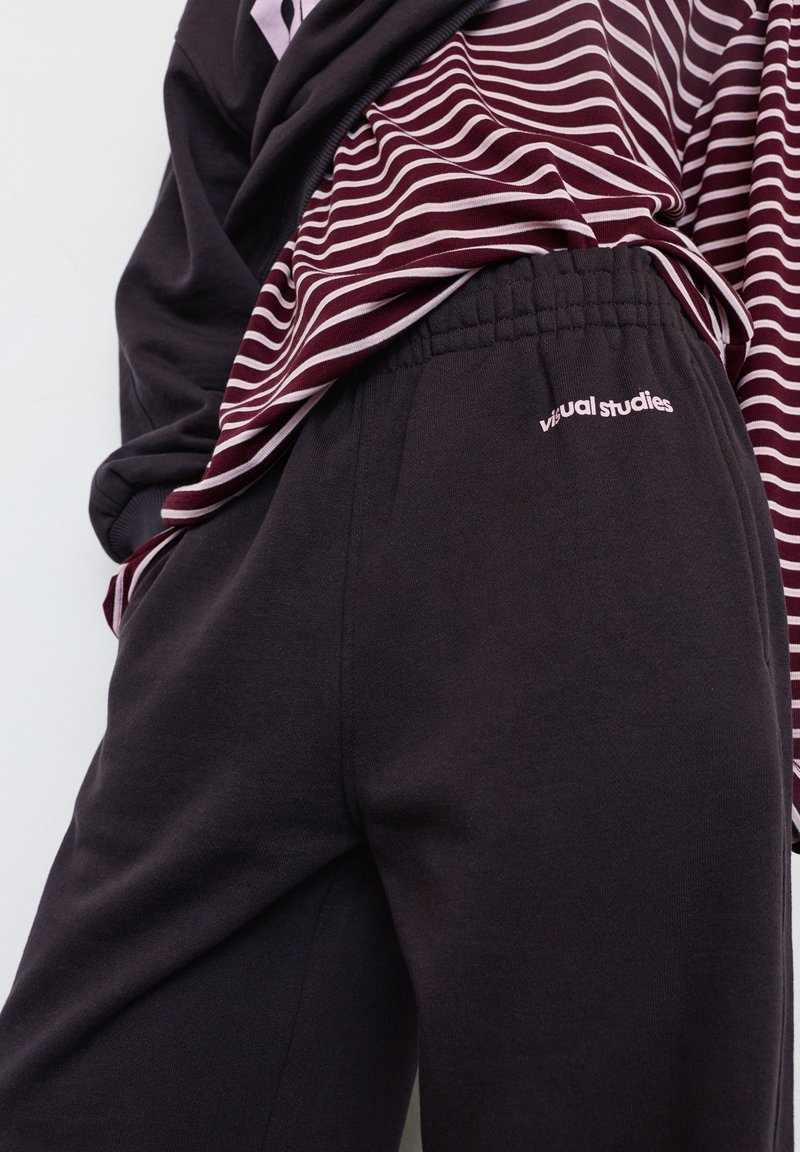 Pantaloni de trening negri cu talie elastică, având un mic logo „studii vizuale”. Purtați cu un top cu dungi burgund și albe.