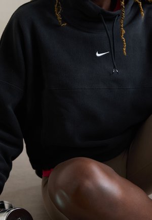 Personne portant un sweat à capuche Nike noir avec des cordons et un pantalon beige, assise avec un genou visible plié, et un haltère argenté à proximité.