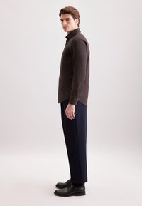 Camicia marrone con bottoni, maniche lunghe, abbinata a pantaloni blu navy e scarpe scure. La camicia presenta una vestibilità su misura e una superficie liscia.