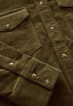 Olivgrön kordflöjlsskjorta med två bröstfickor, silverfärgade tryckknappar och textilstruktur. Närbild av detaljer och sömnad.