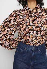 Blusa floreale nera con maniche a sbuffo, caratterizzata da motivi arancioni, rosa e bianchi. Abbinata a jeans in denim scuro a vita alta.