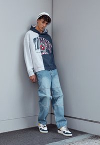 Grå og marineblå hettegensere med "TOMMY" grafikk, avslappede blå jeans og hvite sneakers med blå og gule detaljer, satt mot en nøytral bakgrunn.