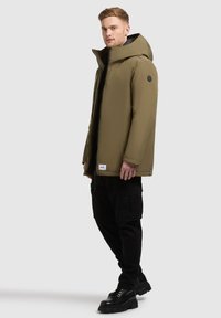 Olivgrön vadderad jacka med stor huva, dragkedjestängning och en logotyp på axeln. Svarta cargobyxor och kraftiga svarta stövlar.