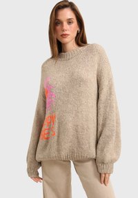 Pull oversized beige avec une texture en tricot épais, ornée d'un graphique vibrant en rose et orange sur le devant.