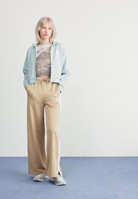 Veste zippée bleu clair sur un crop top graphique, assortie à un pantalon large beige avec des fermetures éclair sur les côtés et des accents blancs. Chaussures à semelle en caoutchouc.