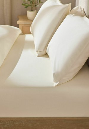 Bett mit cremefarbenem Spannbettlaken und zwei passenden Kissen, neben einem hölzernen Nachttisch mit einer Topfpflanze.
