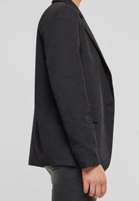 Czarna marynarka o dopasowanym kroju, z pojedynczym zapięciem na guzik, klapami w kształcie litery V i bocznymi kieszeniami. Gładka tekstura, usztywniona konstrukcja i rękawy o średniej długości.