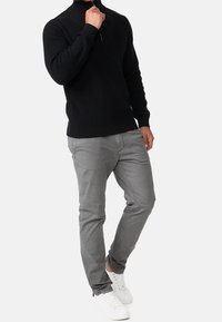 Homme portant un pull en maille noir à demi-fermeture éclair, pantalon gris et baskets blanches, debout devant un fond clair uni.