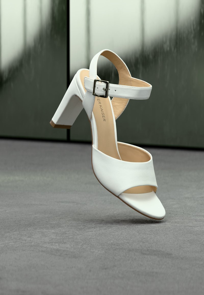 Ανοιχτή γυναικεία σανδάλι σε λευκό χρώμα με ψηλό τακούνι, λουράκι αστραγάλου και ασημένια αγκράφα, σε γκρι μοκέτα με θαμπό φόντο.