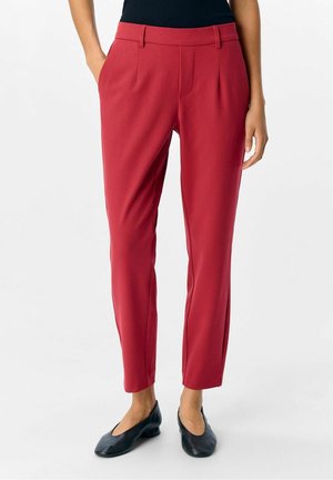 Mujer con pantalones rojos ajustados de talle alto con bolsillos laterales y zapatos planos negros, de pie frente a un fondo blanco liso.